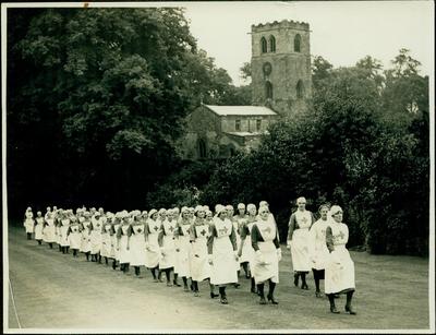 Photograph of a group of female VADs in indoor uniform marching through a field, past a church.