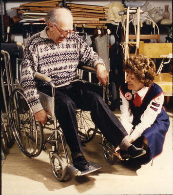 Colour photograph. Elderly man being fitted with a wheelchair in a medical loan depot by a female member of London Branch