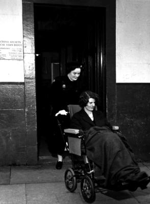 Black and white photograph. A VAD pushing a woman in a wheelchair from the medical loan depot in Oban