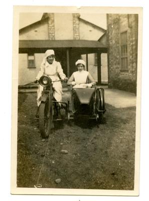 VAD [Dorothy Hancock] in nursing uniform riding motorcycle with another nurse sitting in the sidecar at [Southmead Hospital, Bristol]