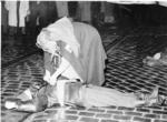 Black and white photograph. Red Cross nurse wearing gas mask and a cloak with a Red Cross emblem tending to a 'patient'