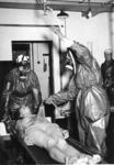 Black and white photograph. Two members dressed in protective clothing and gas masks 'showering' [decontaminating] another member