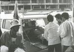 Red Cross medical personnel wearing white coats putting wounded into a Red Cross vehicle with a Red Cross flag for evacuation