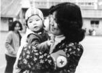 Black and white photograph. Female member of the Hong Kong Red Cross holding up a baby in a refugee camp for Vietnamese boat people
