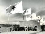 Black and white photograph. Emergency relief houses in Cheskin, Iran. Opening ceremony, view of building with crowd of people standing in front, line of flags in foreground.